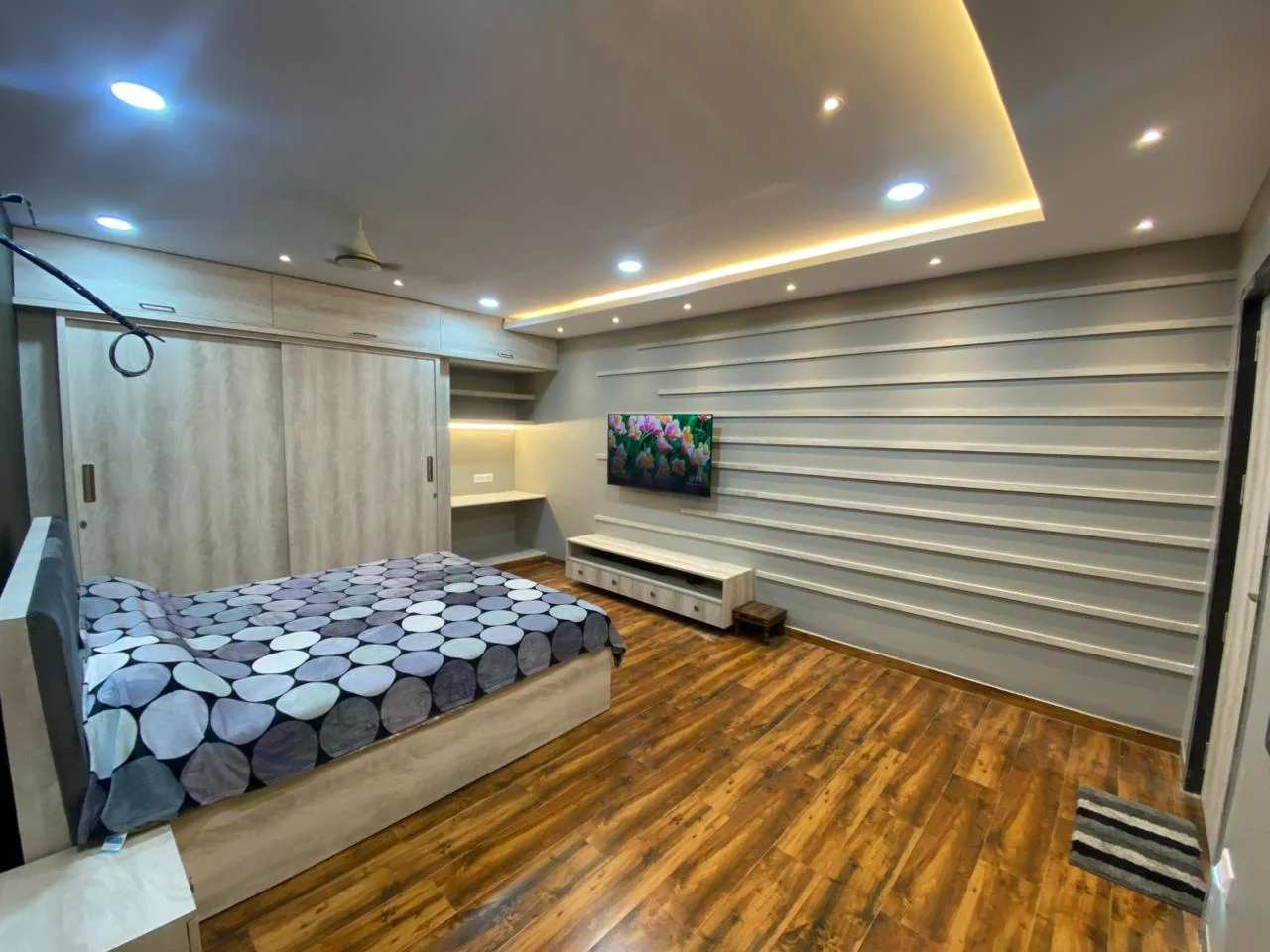 Wide view of a bedroom with slat panelling, a pale laminate wardrobe run and a wall-mounted television